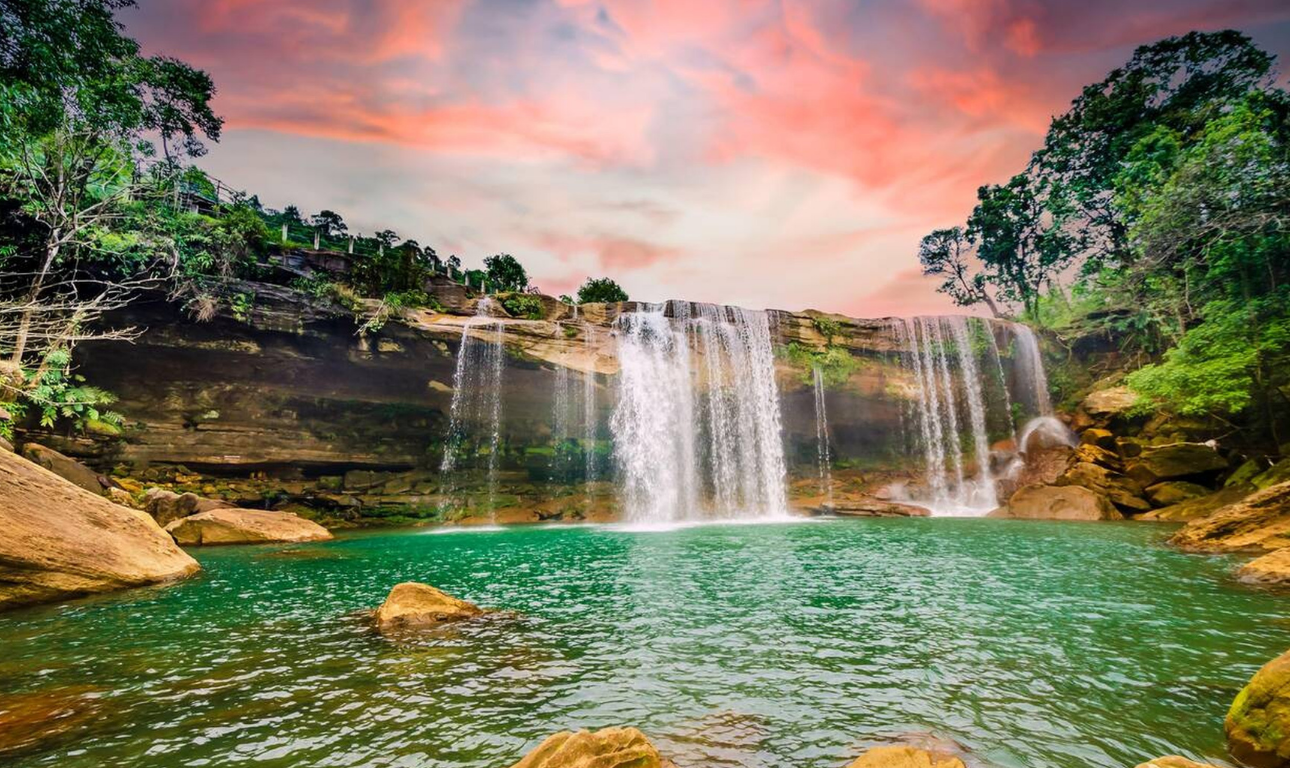 Beautiful natural waterfall flowing into a turquoise pool surrounded by rocks, lush green trees, and a colorful sunset sky.