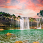 Beautiful natural waterfall flowing into a turquoise pool surrounded by rocks, lush green trees, and a colorful sunset sky.