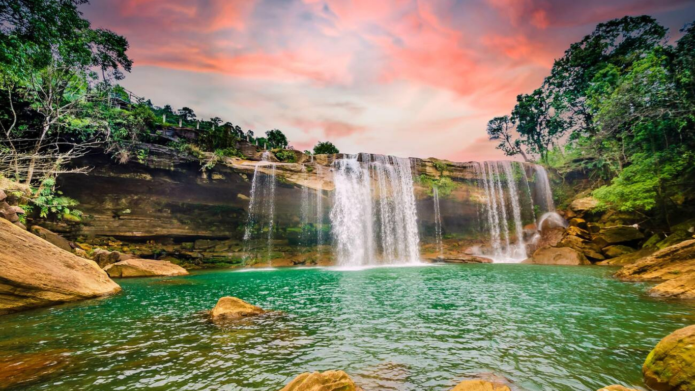 Beautiful natural waterfall flowing into a turquoise pool surrounded by rocks, lush green trees, and a colorful sunset sky.