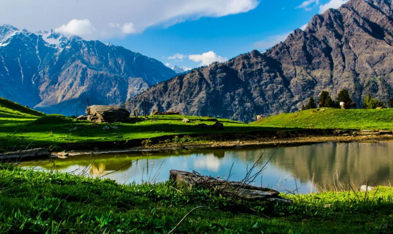 Scenic mountain landscape in Kasol with green meadows, a calm reflective lake, and towering Himalayan peaks under a bright blue sky.