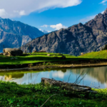 Scenic mountain landscape in Kasol with green meadows, a calm reflective lake, and towering Himalayan peaks under a bright blue sky.