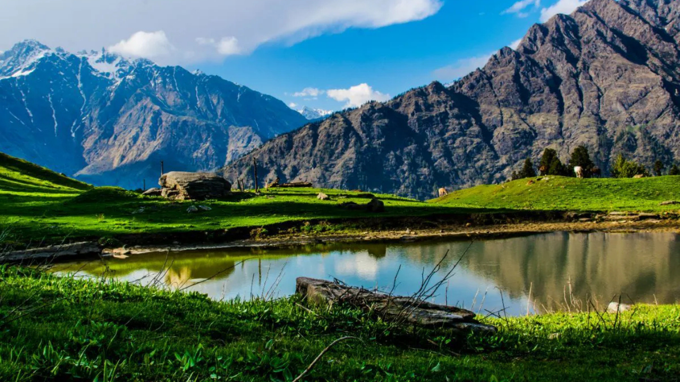Scenic mountain landscape in Kasol with green meadows, a calm reflective lake, and towering Himalayan peaks under a bright blue sky.