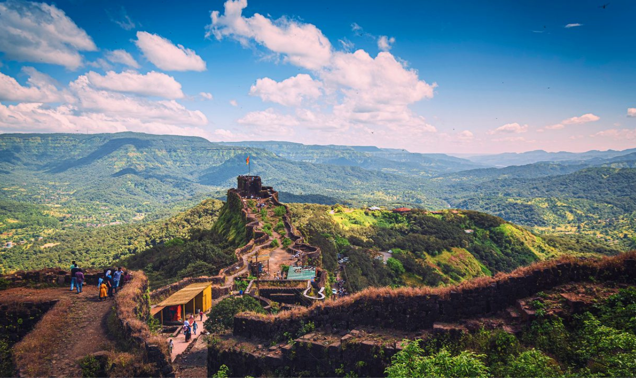 Scenic view of Mahabaleshwar hills with historic mountain fort, lush green valleys, winding pathways, and a bright blue sky with clouds.