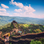 Scenic view of Mahabaleshwar hills with historic mountain fort, lush green valleys, winding pathways, and a bright blue sky with clouds.