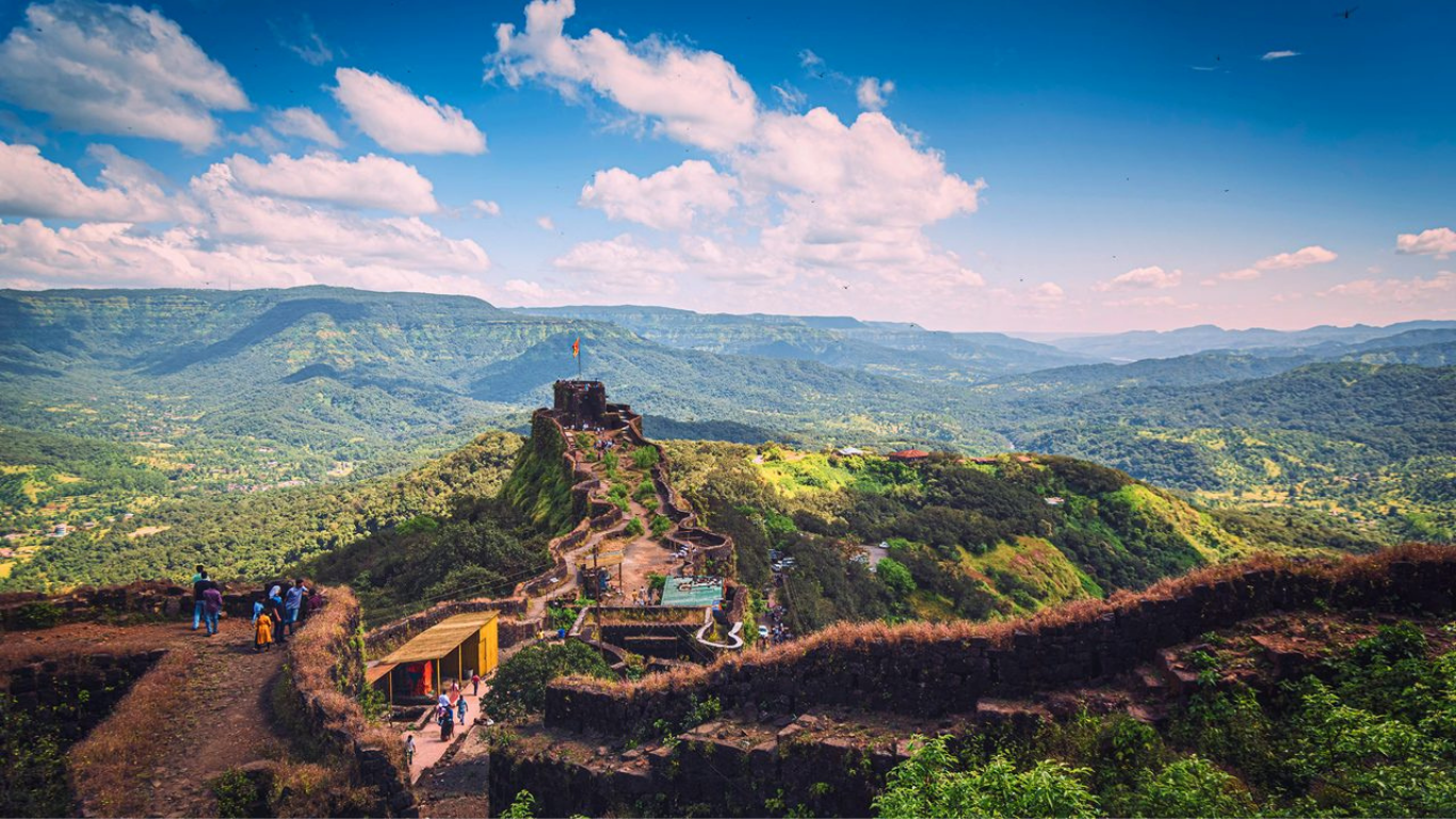 Scenic view of Mahabaleshwar hills with historic mountain fort, lush green valleys, winding pathways, and a bright blue sky with clouds.