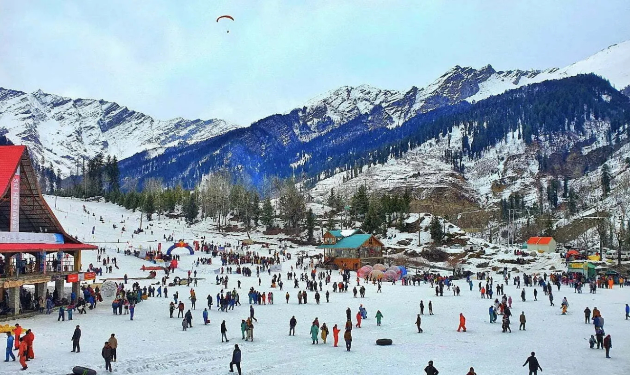 Snow-covered landscape in Manali with tourists enjoying winter activities, surrounded by pine trees and majestic Himalayan mountains.