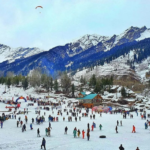 Snow-covered landscape in Manali with tourists enjoying winter activities, surrounded by pine trees and majestic Himalayan mountains.