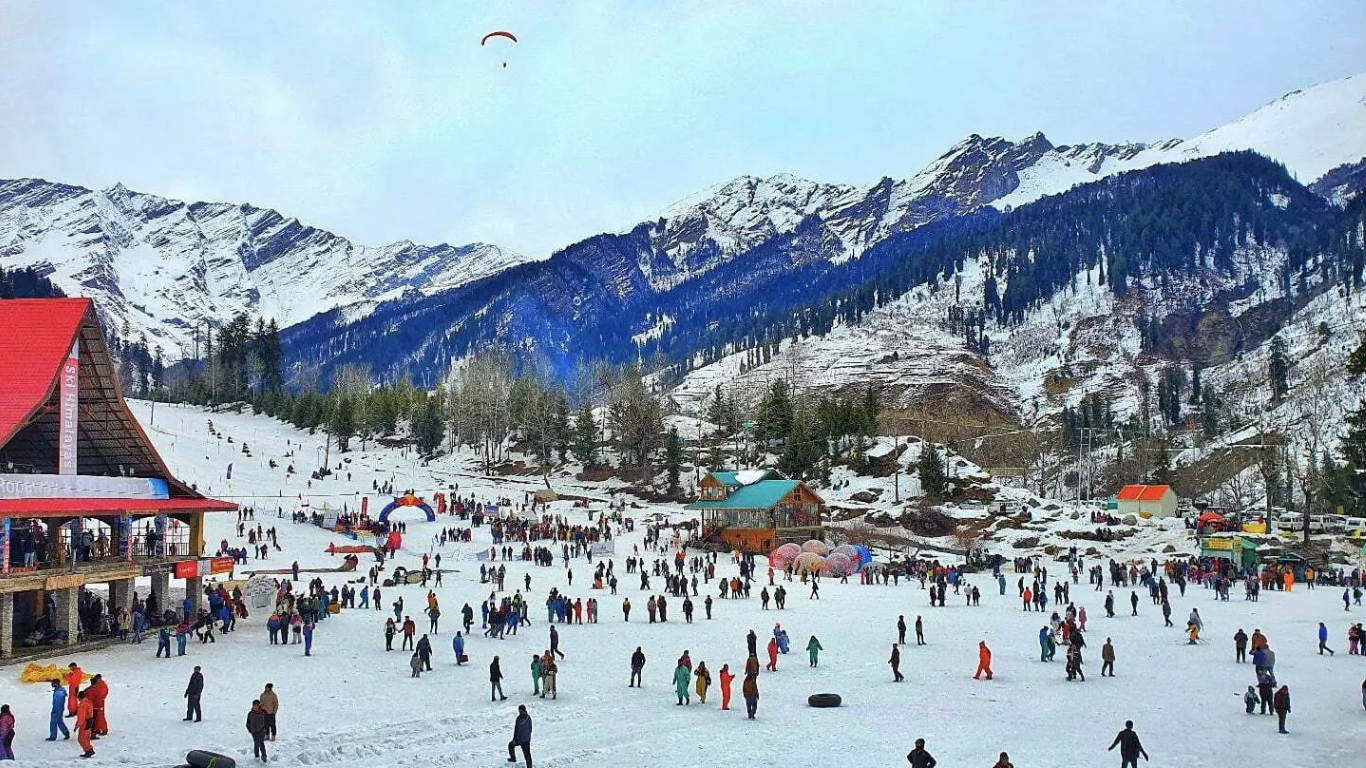 Snow-covered landscape in Manali with tourists enjoying winter activities, surrounded by pine trees and majestic Himalayan mountains.