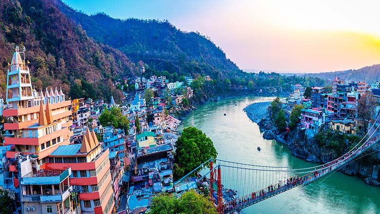 Beautiful view of Rishikesh featuring the Ganga River, suspension bridge, colorful buildings, and scenic hills during sunset.